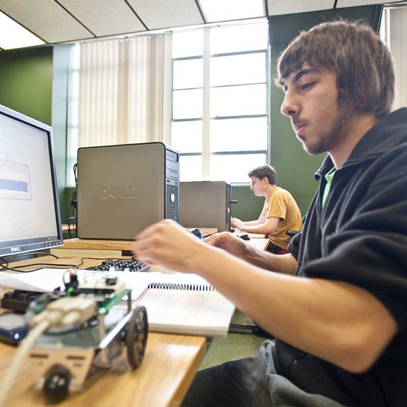 A UMass Lowell computer engineering student works at a computer with a small robot nearby 