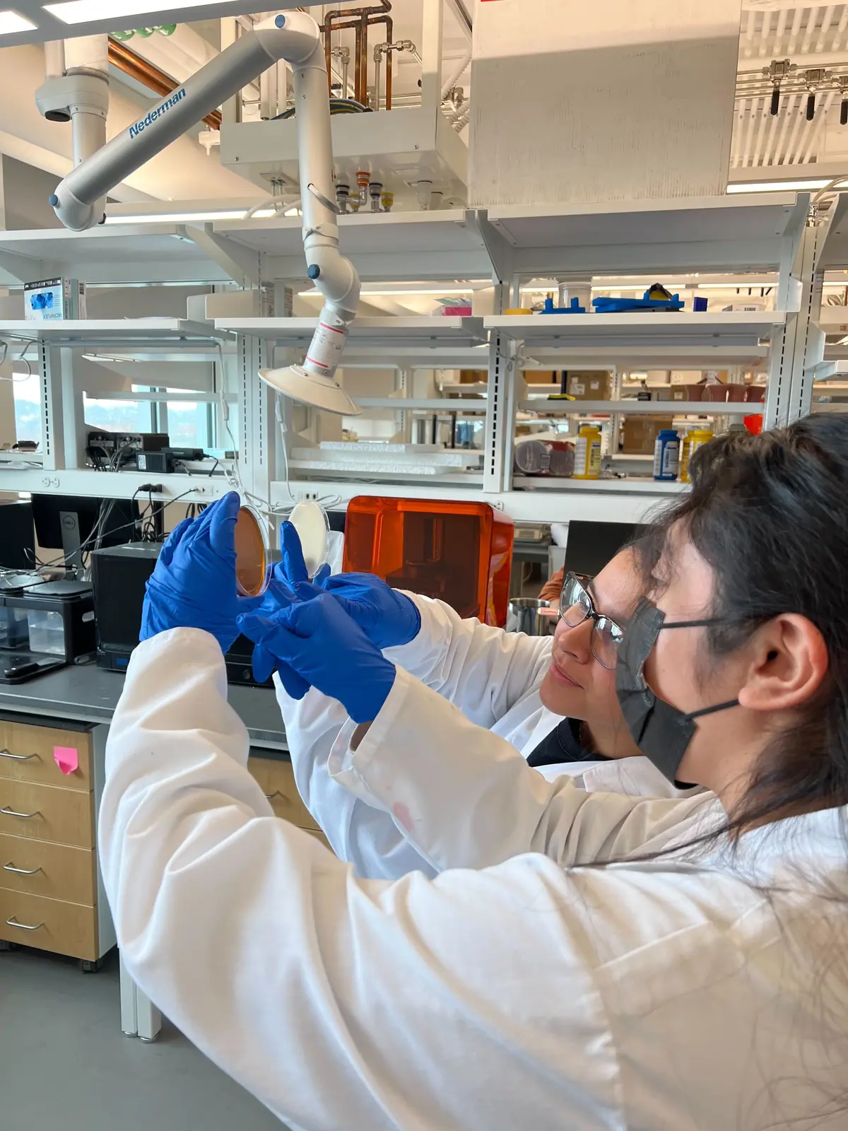 Two women in lab coats and gloves work in a lab