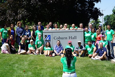 A woman takes a group photo of people in green T-shirts gathered around a sign for Coburn Hall
