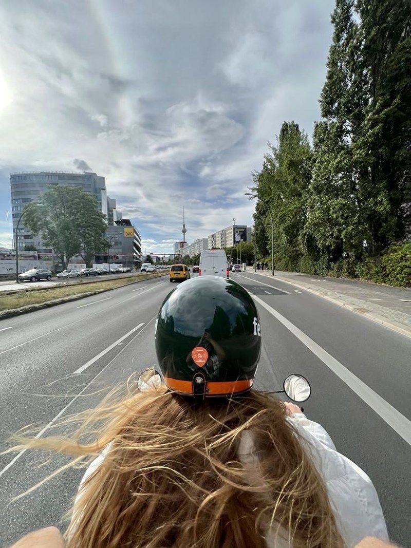 A photograph taken from the back seat of a moped on a city highway that shows the driver's black helmet and long hair moving in the wind on a bright, cloudy afternoon.