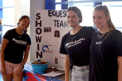 Three women in black shirts pose for a photo next to a display for their equestrian team