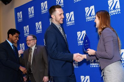 A man shakes hands with a woman who is holding an award while two other men talk in the background