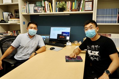 Two men wearing face coverings pose for a photo at a desk in front of a computer monitor