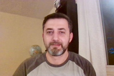 A man with short hair and a bear poses for a photo while on a video chat from home