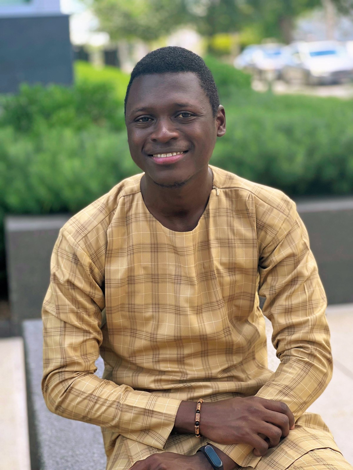 Esan Olorunfemi James sitting down outside  wearing a yellow plaid shirt and smiling at the camera. 