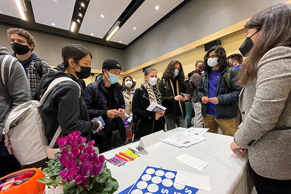 A half dozen students wearing face coverings gather around a table, which has flowers, pens and papers on it