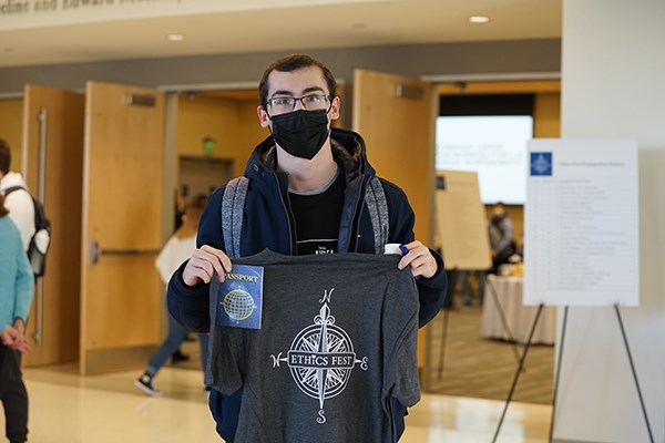 A male student in glasses and a face covering holds a T-shirt and small booklet while posing for a photo
