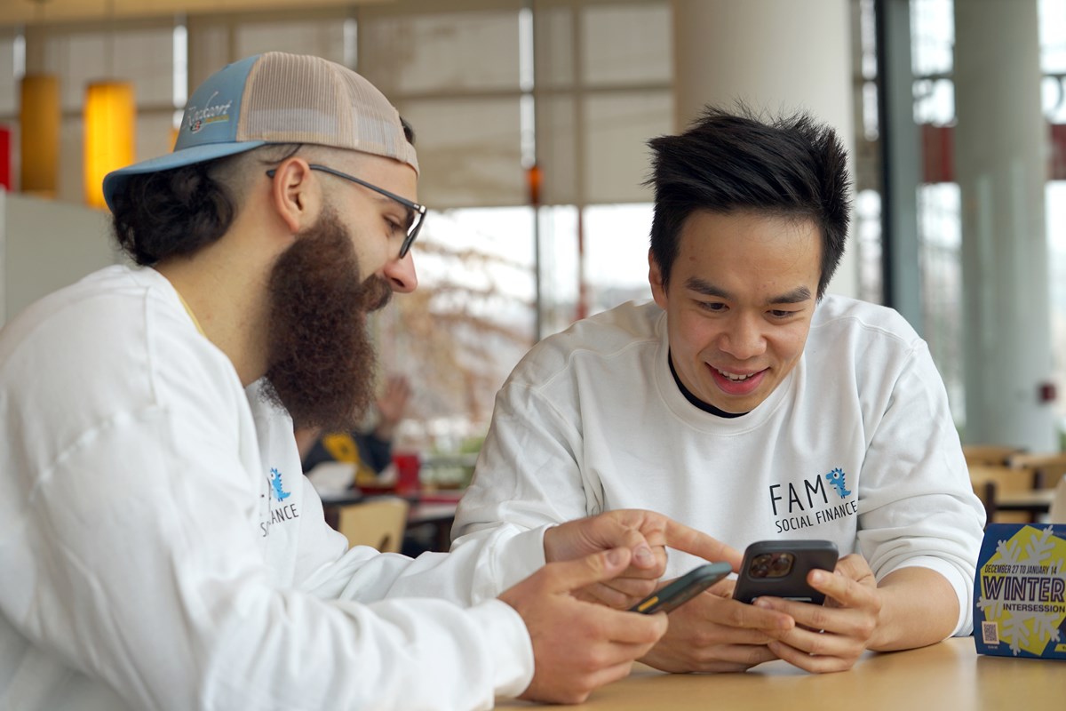Two young men in white shirts look at their phones while sitting at a table.