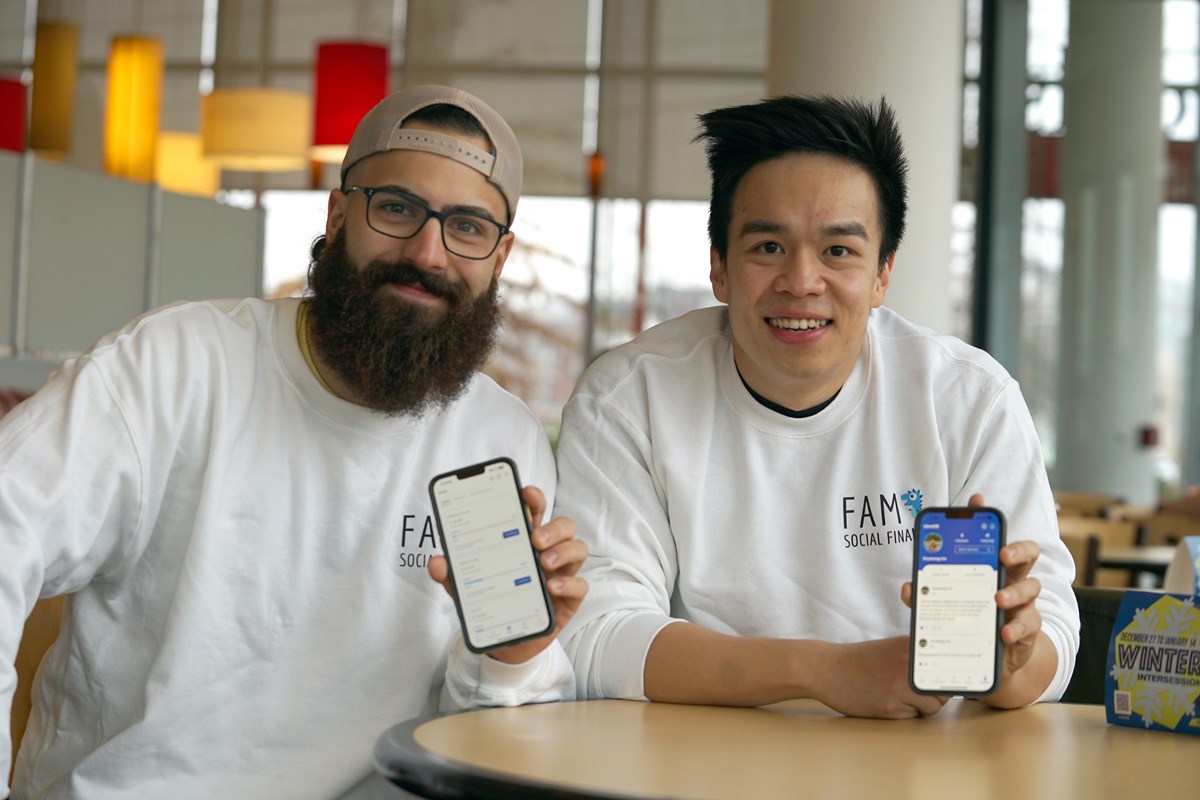 A man with a beard and another man with dark hair pose for a photo while holding their phones.