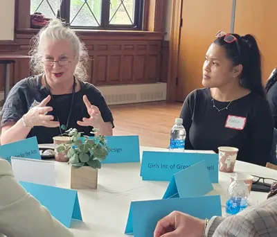 Woman speaks to other woman with Girls Inc signs on round table