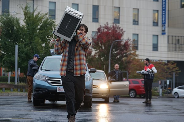 A student carries an air conditioner on his shoulder in a parking lot while another student collects recycled items from a car in the background