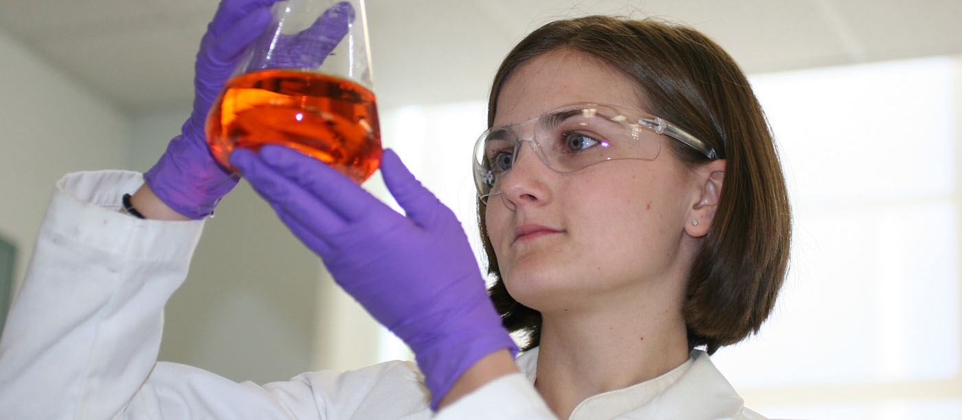 young woman wearing safety glasses and rubber gloves holding up a glass beaker containing orange liquid