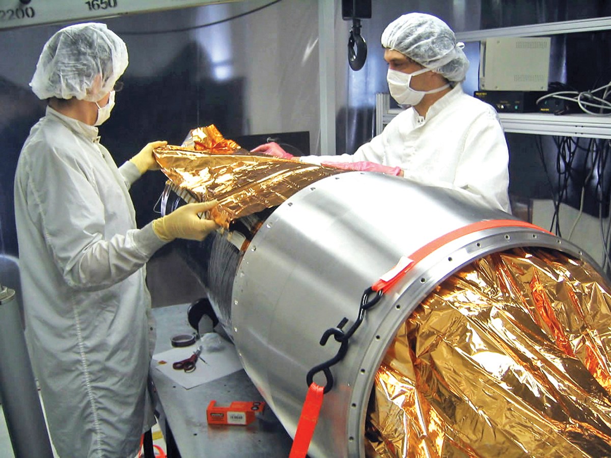Two people in lab coats and caps placing a gold material on a Space Imaging Instrument.