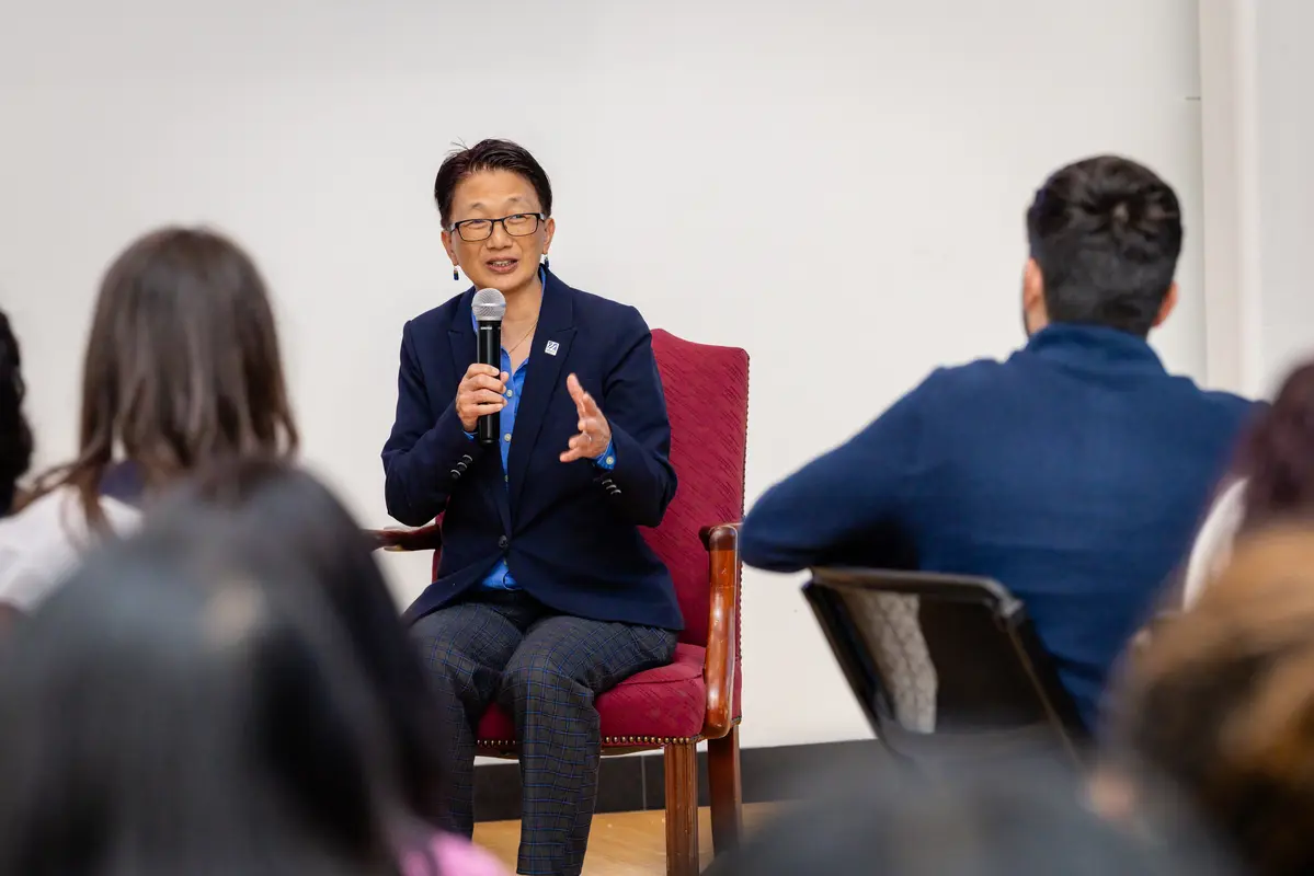 A seated woman wearing glasses gestures while speaking with a microphone to students.