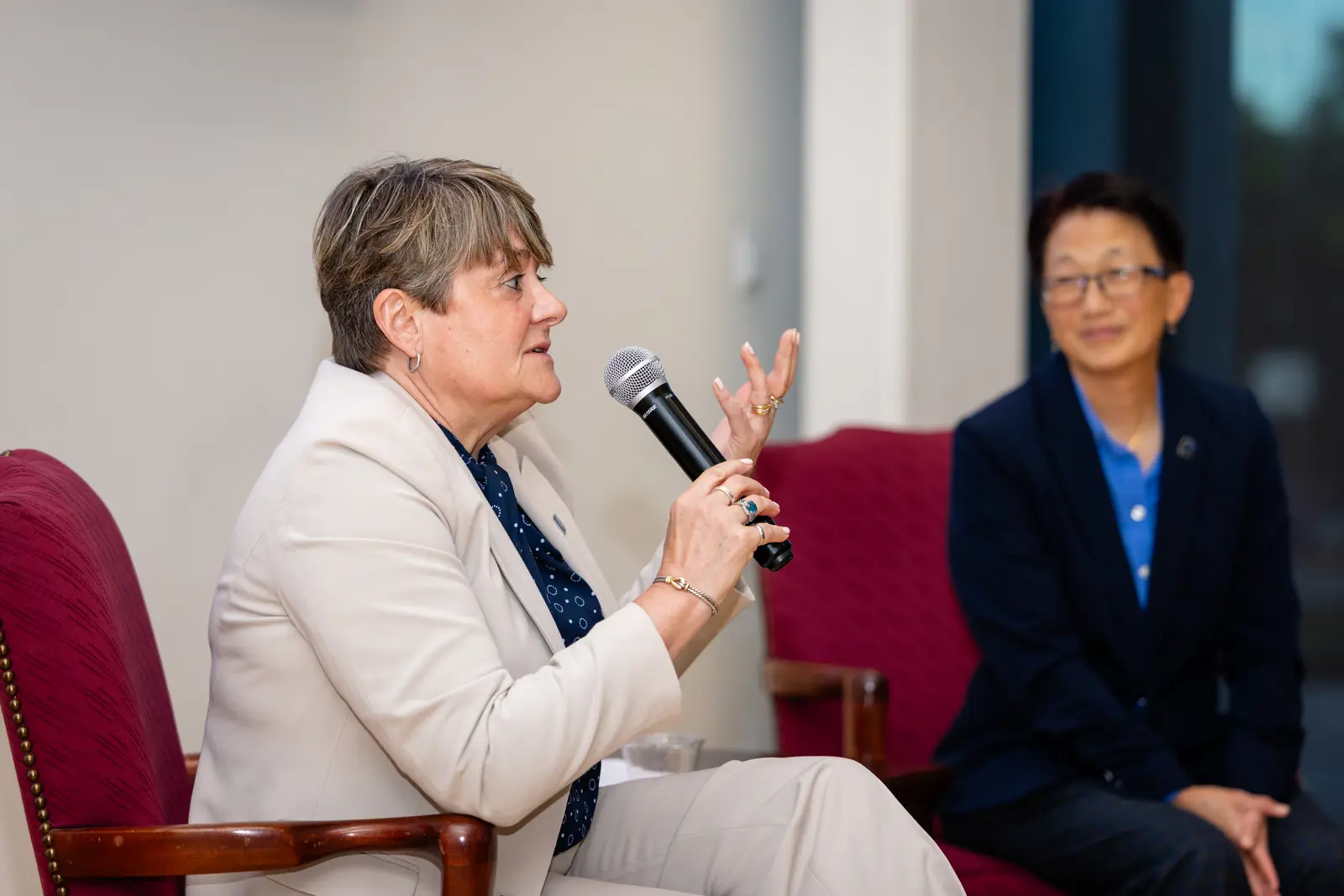 A seated woman gestures with her left hand while speaking into a microphone while another woman looks on.