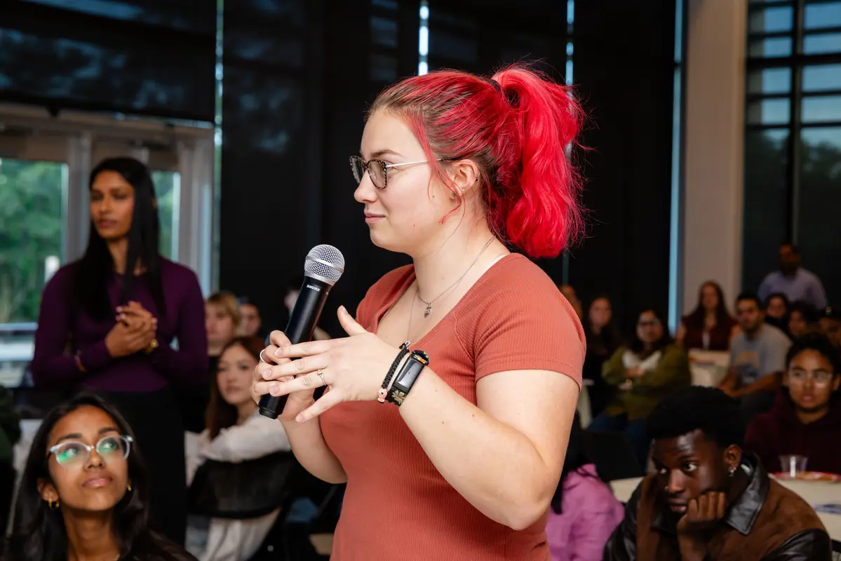 A woman with bright red hair and glasses stands and holds a microphone.