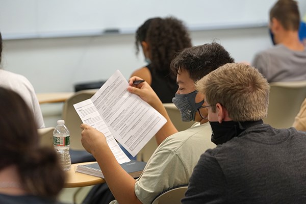 A student holds a paper while another student looks over his shoulder in a classroom
