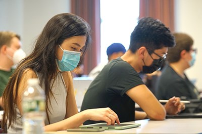 A female student in a face mask types on an ipad while sitting next to a male student at a desk in a classroom