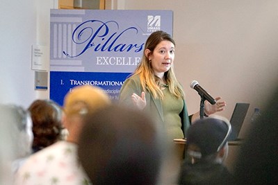 A woman gestures with her hands while speaking into a microphone to people in a room