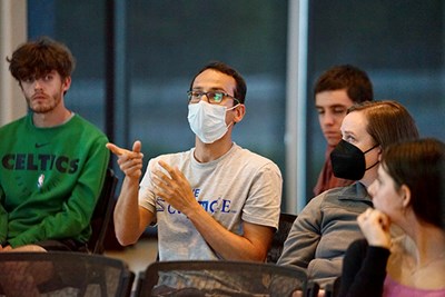 A young man wearing a face covering and glasses asks a question while seated with other people