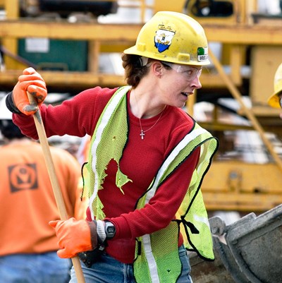 A young woman wearing safety gloves, hard hat and safety vest holds a broom. Future of Work photos by Paul Shoul woman construction