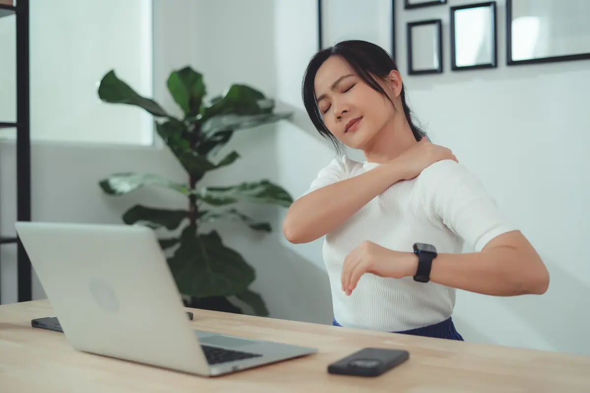 A woman rubs her shoulder with her eyes closed while seated at a table working on a laptop. 