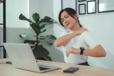 A woman rubs her shoulder with her eyes closed while seated at a table working on a laptop. 