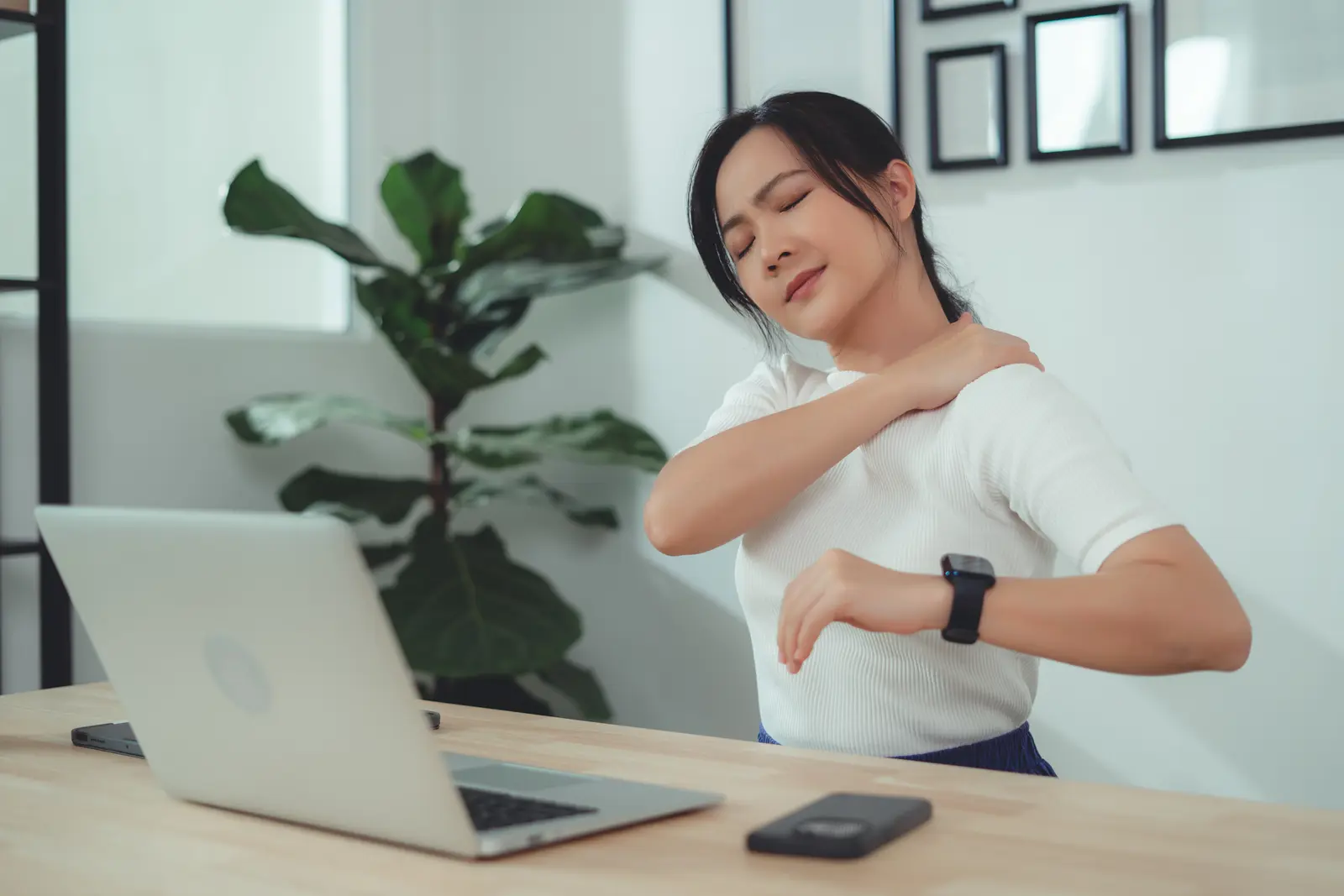 A woman rubs her shoulder with her eyes closed while seated at a table working on a laptop. 