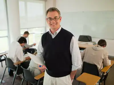A person stands at the front of a classroom while others work at desks in the background.