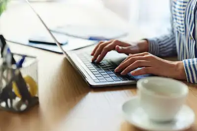 Close up of a person typing on laptop at a desk with a cup on a saucer and a box with pens, tape etc.