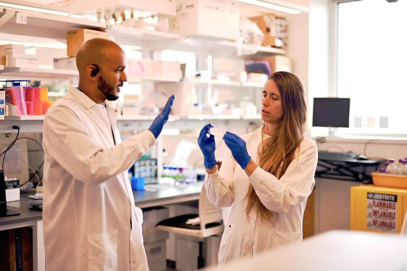 A man and a woman in white lab coats and blue gloves work in a college lab.