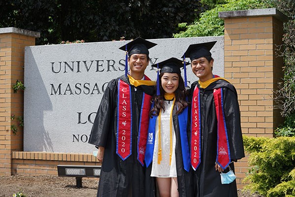 Two male Asian students flank a female Asian student in the regalia in front of North Campus sign