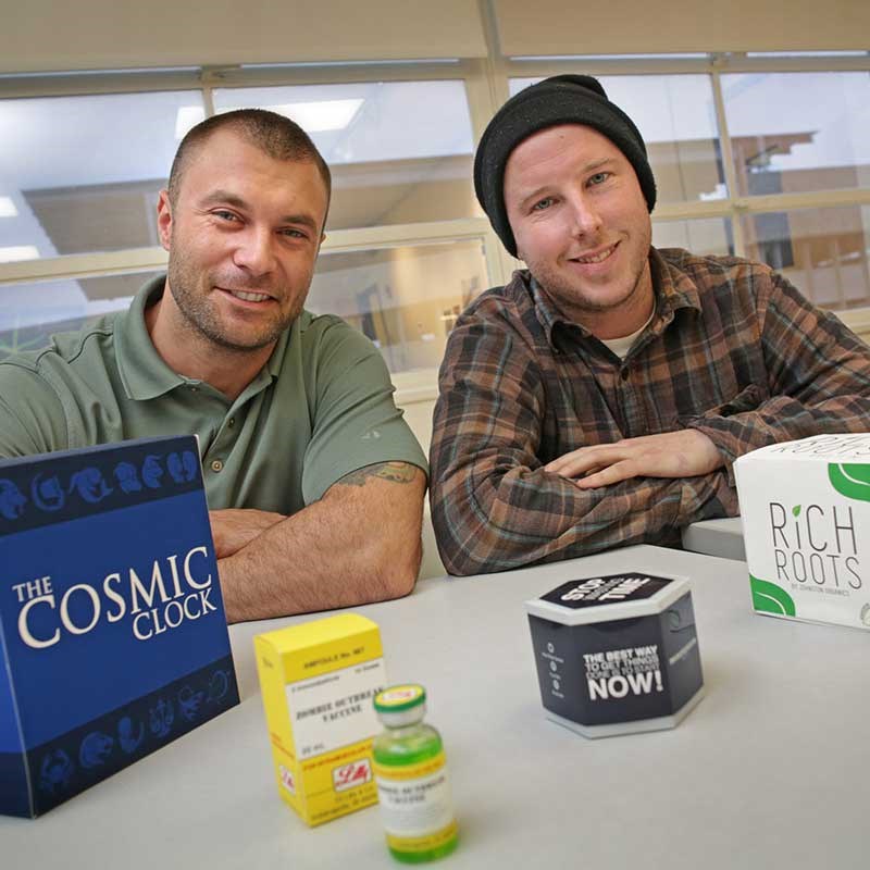 Two graphic design students seated next to their packaging designs in a UMass Lowell studio