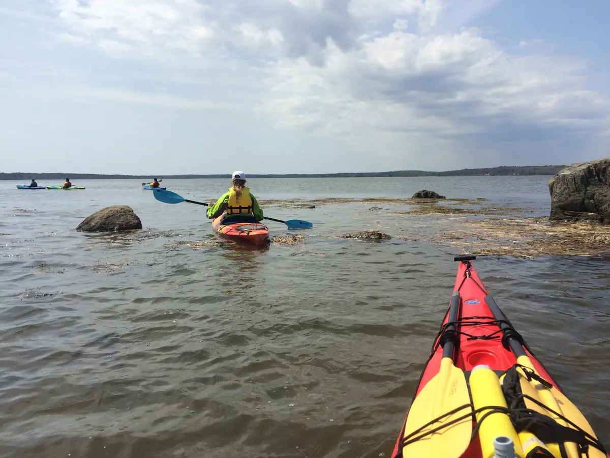 The bow or front of a photographers red sea kayak sits at the bottom right of the image with rescue gear atop. A few kayakers can be seen sporting various styles and colors of kayaks off in the distance.