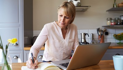Woman writing on paper while working on laptop in her kitchen