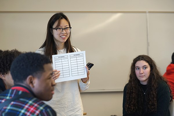 A woman in glasses smiles while holding up a piece of paper as seated students look on