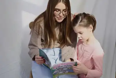 A young woman in glasses reads a childrens book to a young girl in a pink shirt.