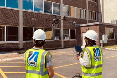 Two researchers wearing UMass Lowell vests watch a drone fly.