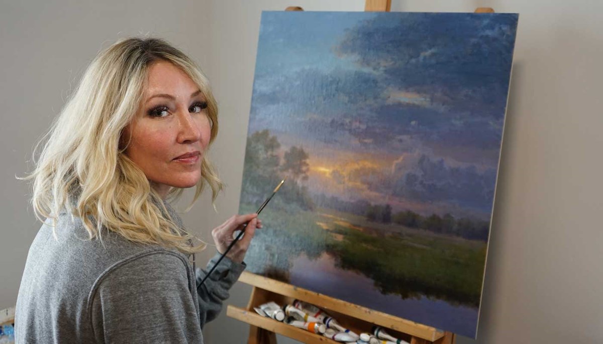Hillary Scott holding a paintbrush in front of a landscape painting