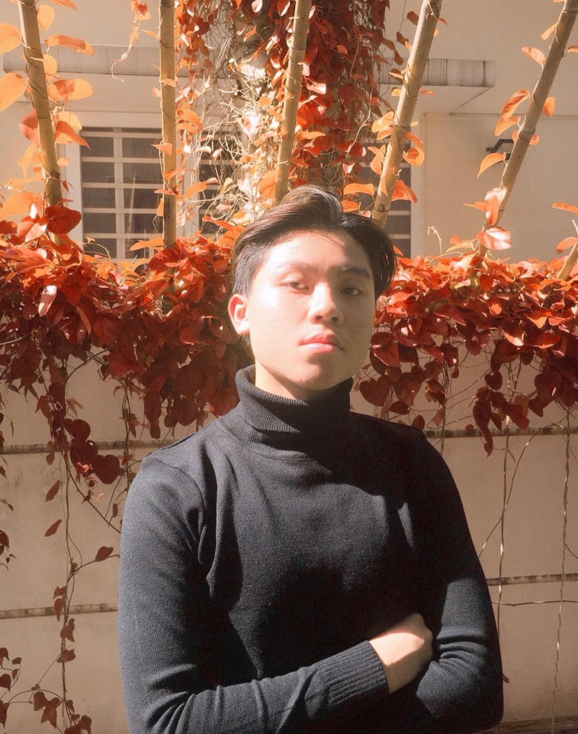 Hoang Nguyen wearing a black turtleneck standing outside in front of a bunch of leaves looking at the camera.