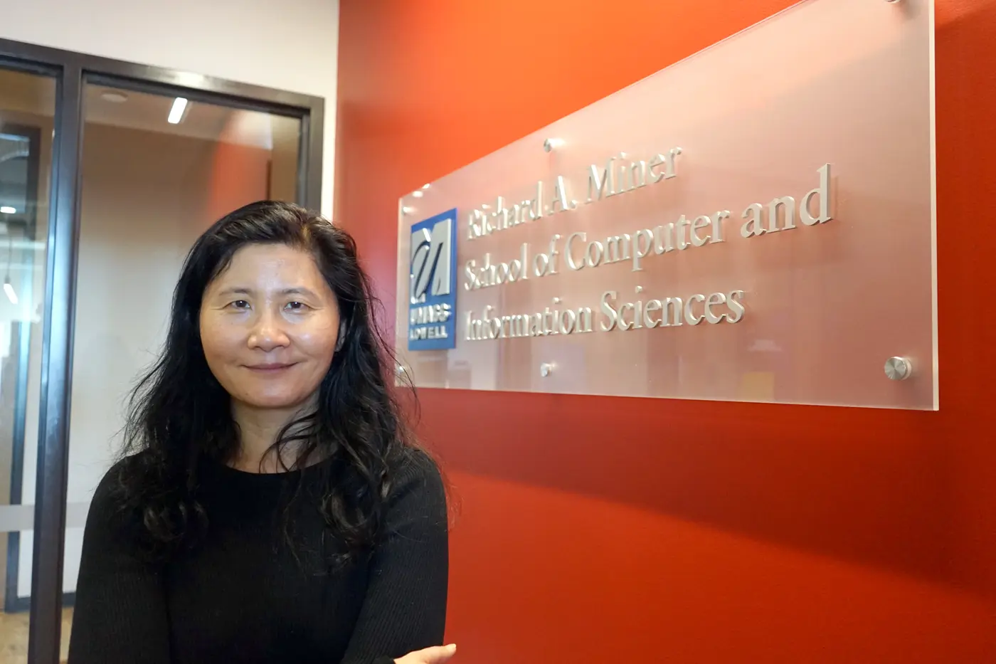 A woman with dark hair poses for a photo next to an orange wall with a glass office sign on it.