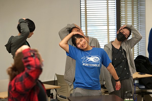 A student smiles while holding her head and stretching with other students in a classroom