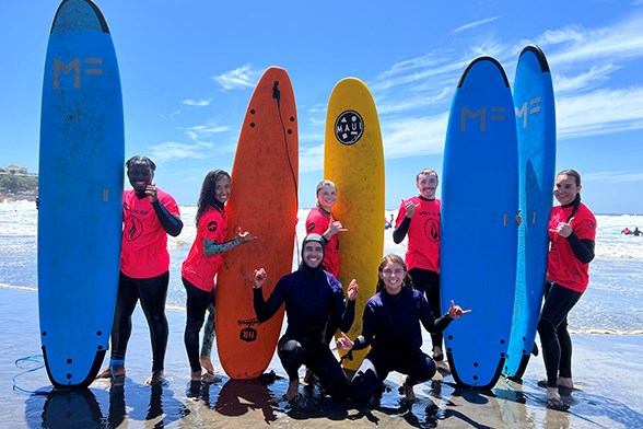 Seven young men and women pose with surf boards on a beach