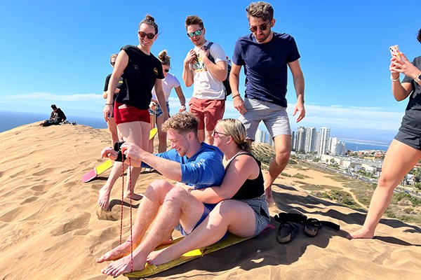 A male and female student prepare to slide down a sand dune while several other students look on