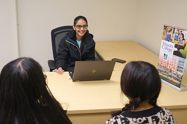 A young woman in glasses smiles while sitting at a desk with a laptop and talking to two people