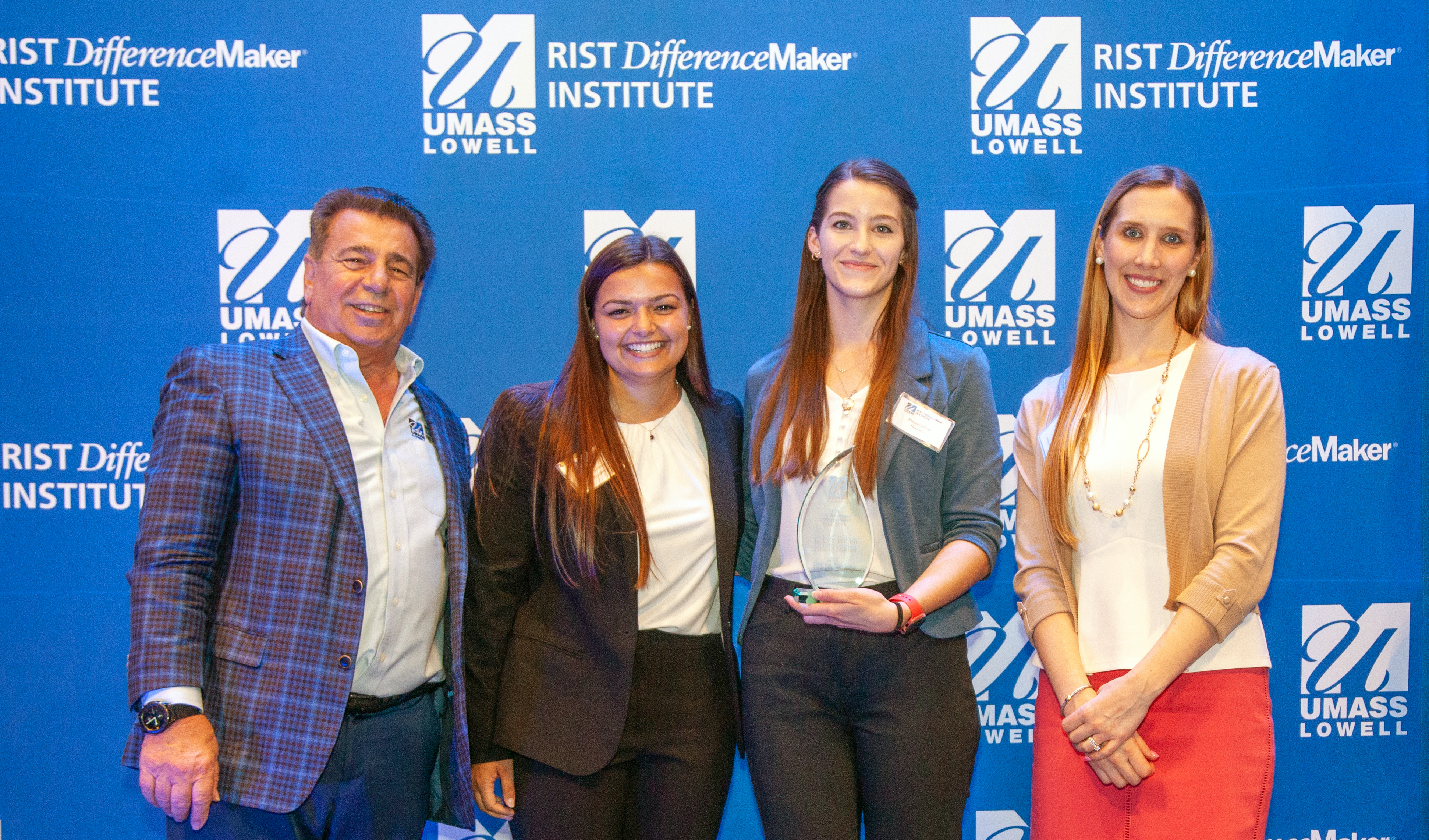 2 female students from the HOPPERS team holding an award pose with Brian Rist and Holly Lalos of Difference Makers against a blue UMass Lowell backdrop.