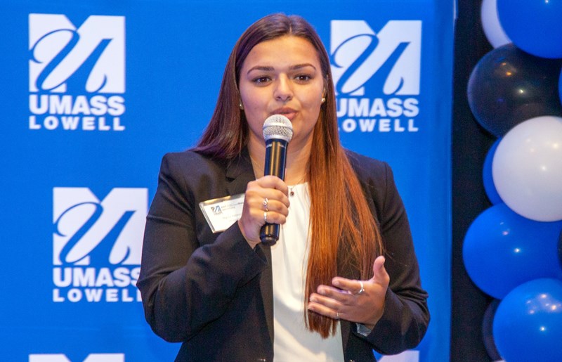 One of the members HOPPERS team holding a microphone and speaking in front of blue UMass Lowell backdrop.