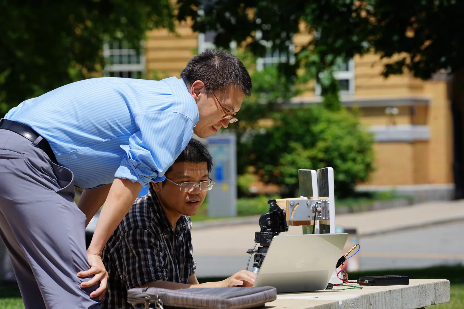 Two men with dark hair and glasses look at scientific instruments set up outside on a college campus.