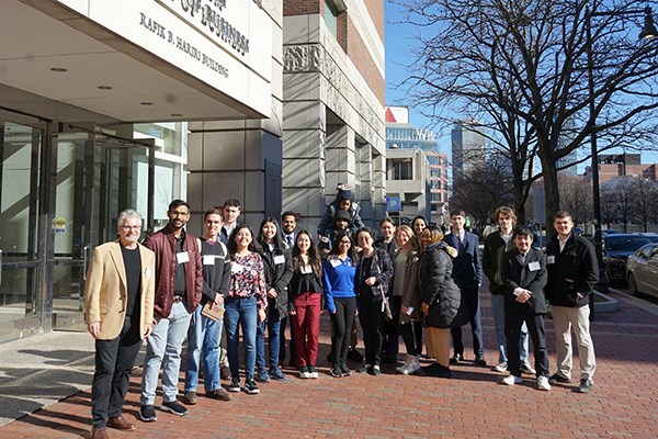 Two dozen men and women pose for a group photo outside in front of a building with Boston in the background