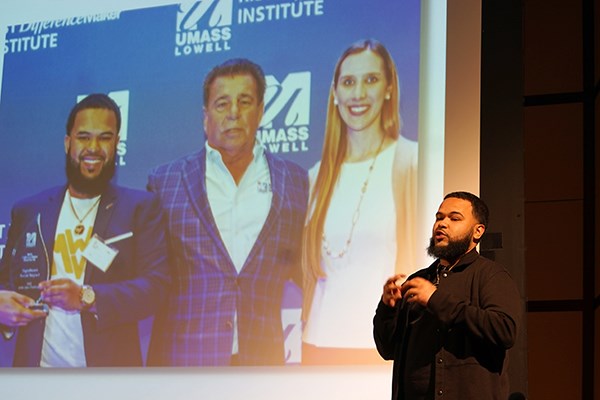 A young man with a beard makes a presentation on a stage in front of a photo of three people
